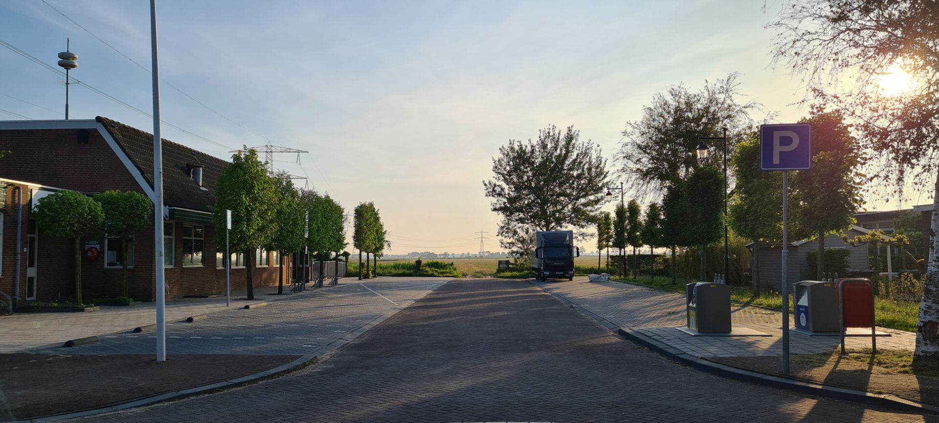 Straatbeeld bij het gebouw: blauw parkeerbord, bomen langs de weg en het polderlandschap in avondlicht.
