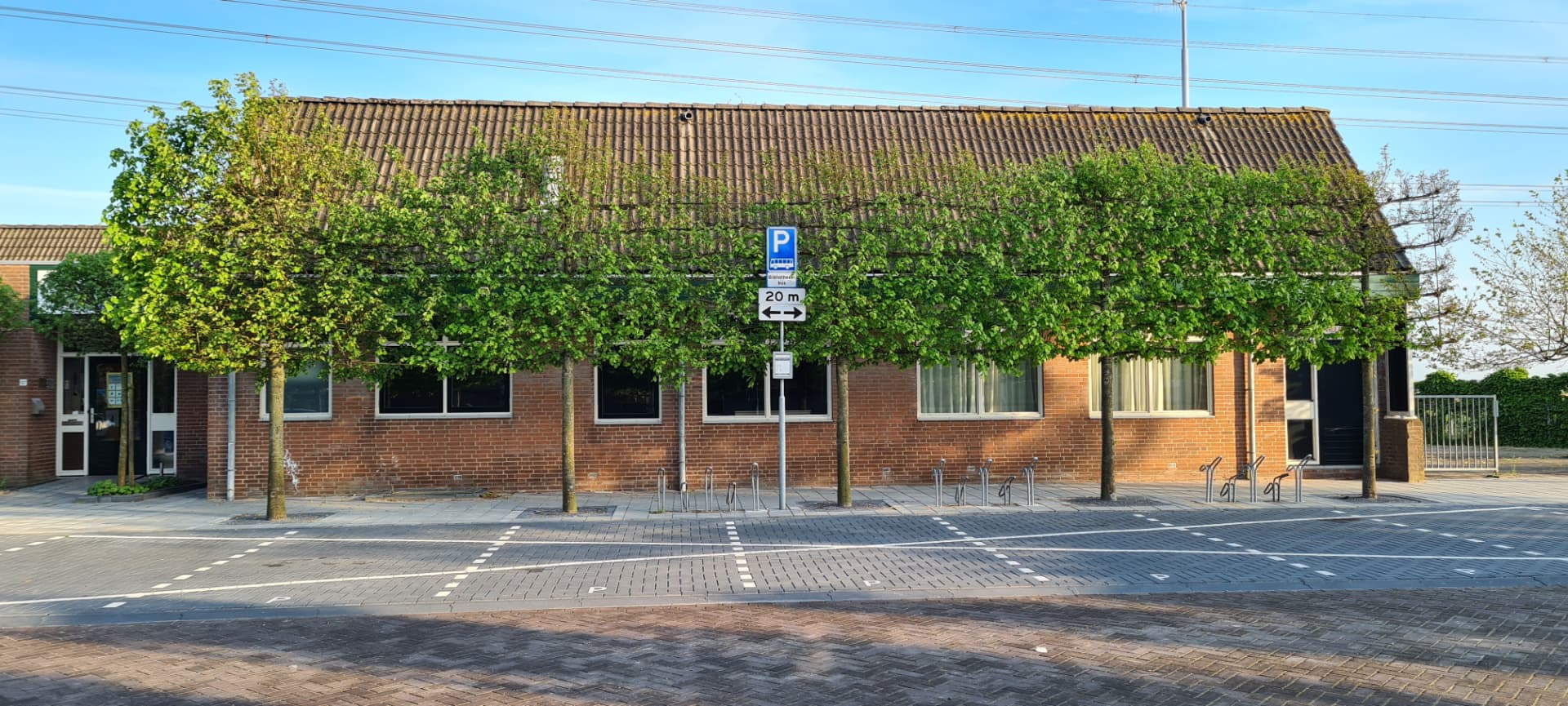 Voorgevel van het bakstenen gebouw met volgroeide bomen, fietsenrekken en parkeerstroken ervoor.