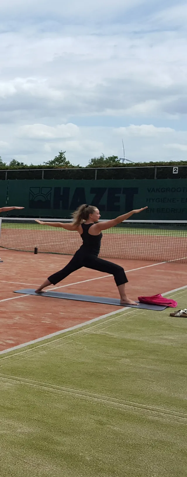 Een persoon staat in een krijgershouding op een yogamat op een gravel tennisbaan, met de armen gespreid.